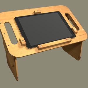 Puede incluir: Un soporte de tableta de madera con una tableta negra apoyada en la parte superior. El soporte tiene un asa incorporada y está diseñado para sostener la tableta en un ángulo para una visualización cómoda.