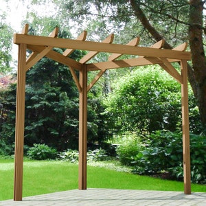 May include: A wooden pergola with a natural brown finish. The pergola has a rectangular shape and is made of sturdy beams. It is standing on a grassy lawn with trees in the background.