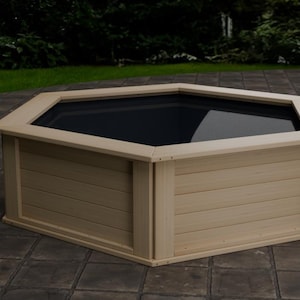 May include: A light-colored, octagonal wooden planter box with a dark interior. The planter has a wide, flat top and is constructed with horizontal wooden planks. The box is set on a stone patio, with greenery in the background.