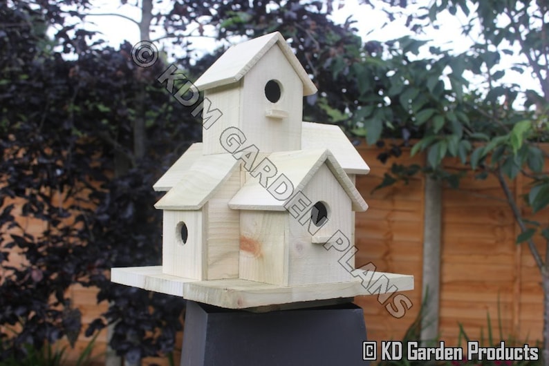 May include: A multi-level wooden birdhouse with multiple compartments and circular entry holes. The birdhouse is constructed from light-colored wood and features a pitched roof design. The birdhouse is mounted on a black stand, with a natural outdoor background.