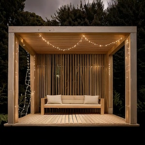 May include: A wooden outdoor structure with a slatted back and roof, featuring a bench with two pillows. String lights are draped across the ceiling and down the side. The structure is set against a dark background of trees.