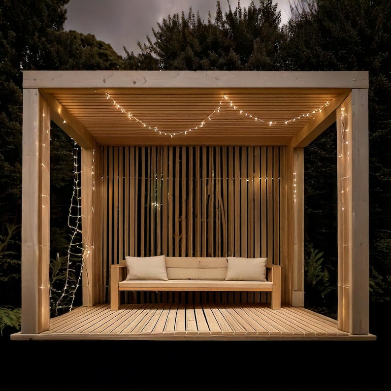 May include: A wooden outdoor structure with a slatted back and roof, featuring a bench with two pillows. String lights are draped across the ceiling and down the side. The structure is set against a dark background of trees.