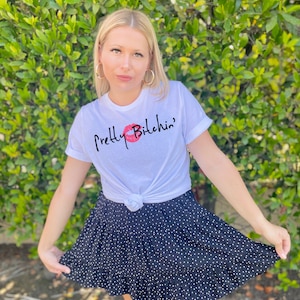 May include: A white t-shirt with the text "Pretty B*tch*n'" and a red lip print. The shirt is tied at the waist and paired with a black skirt with white polka dots.