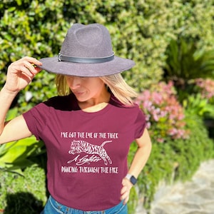 May include: A burgundy t-shirt with white text and a tiger graphic. The text reads "I've got the eye of the tiger, a fighter dancing through the fire." A grey fedora hat with a black band is also visible.