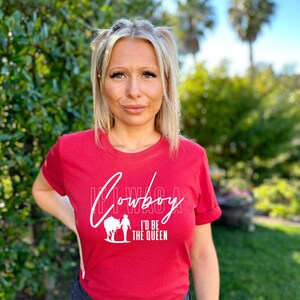 Peut inclure: T-shirt rouge avec un graphisme blanc qui dit "If I Was A Cowboy I'd Be The Queen" et une silhouette de femme &agrave; cheval.