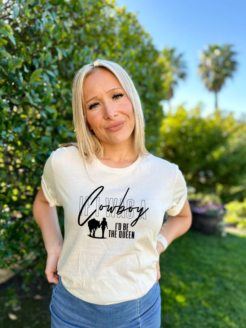 Peut inclure: Une femme portant un t-shirt blanc avec du texte noir qui dit "If I Was A Cowboy I'd Be The Queen" et une silhouette de cow-boy &agrave; cheval. Elle porte un short en jean bleu et a les cheveux blonds.