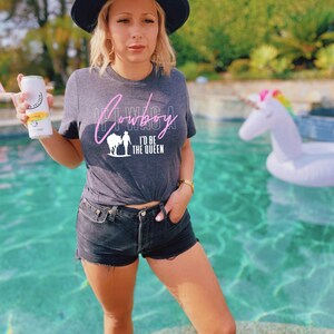 Peut inclure: Une femme portant un t-shirt gris avec le texte "If I Was A Cowboy I'd Be The Queen" en lettres roses. Elle porte un chapeau noir et un short en jean. Elle se tient au bord d'une piscine avec un flotteur en forme de licorne en arri&egrave;re-plan.