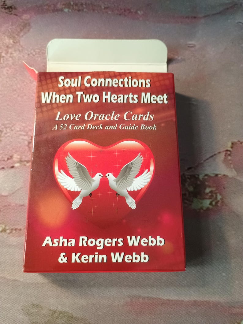 May include: A red box with a white border and a white heart with two white doves inside. The text on the box reads "Soul Connections When Two Hearts Meet Love Oracle Cards A 52 Card Deck and Guide Book Asha Rogers Webb & Kerin Webb".