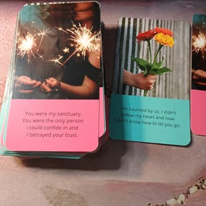 May include: Two cards with pink and blue borders and text. The first card shows a couple holding sparklers and the text "You were my sanctuary. You were the only person I could confide in and I betrayed your trust.". The second card shows a hand holding a bouquet of orange and red flowers and the text "I'm haunted by us. I didn't follow my heart and now I don't know how to let you go."