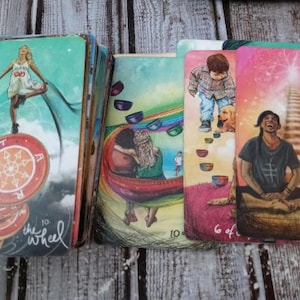 May include: A tarot card reading with ten cards laid out on a wooden surface. The cards are illustrated with colourful images and text. The cards include "The Wheel", "6 of Cups", and "5 of the Hierophant".