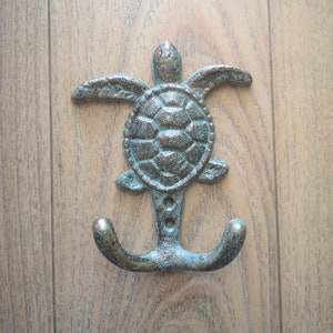 May include: A decorative metal hook shaped like a sea turtle with a double hook at the bottom. The hook is painted in a light blue and green color.