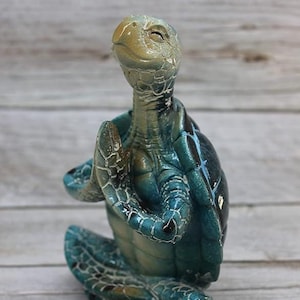 May include: A blue and green ceramic turtle figurine in a yoga pose with its hands folded in front of its chest.