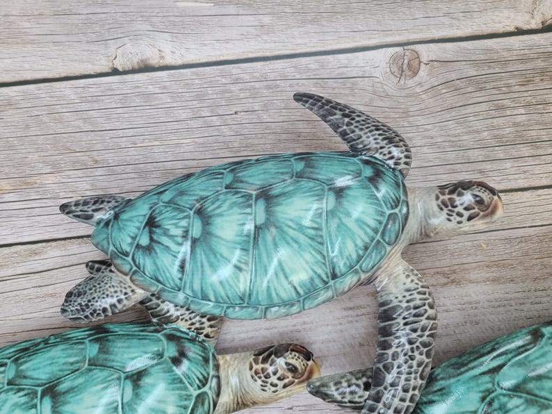 May include: Several decorative sea turtles with teal shells and detailed patterns. The turtles have textured skin and are arranged on a light wood-grain surface. The turtles are a decorative item.