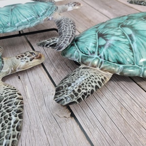 May include: Three decorative sea turtles with teal and green shells and patterned flippers are arranged on a light wood surface. The turtles have detailed textures and varying sizes, creating a coastal theme.