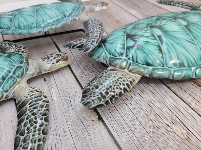 May include: Three decorative sea turtles with teal and green shells and patterned flippers are arranged on a light wood surface. The turtles have detailed textures and varying sizes, creating a coastal theme.