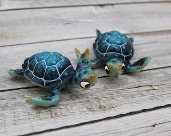 Peut inclure: Deux figurines de tortues marines en céramique. Les tortues ont des carapaces bleues avec un motif blanc et des accents vert clair et jaune. Elles sont posées sur une surface en bois clair vieilli.
