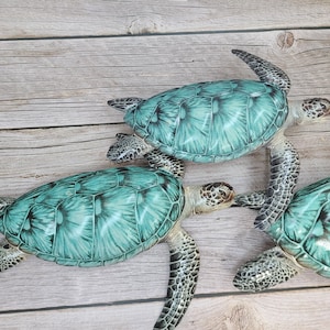 May include: Three decorative sea turtles with turquoise shells and detailed patterns. The turtles are arranged on a light brown wooden surface, showcasing their realistic design and color variations. The turtles' flippers and heads are visible.