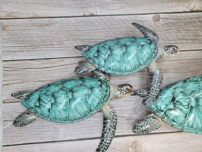 May include: Three decorative sea turtles with turquoise shells and detailed patterns. The turtles are arranged on a light brown wooden surface, showcasing their realistic design and color variations. The turtles' flippers and heads are visible.