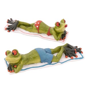 May include: Two ceramic frog figurines sunbathing on striped towels. One frog wears a red bikini with white polka dots, the other wears blue swim trunks. Both have red eyes, black hands and feet, and are lying down.