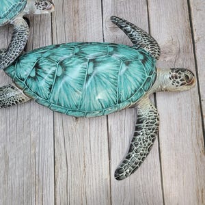May include: Several decorative sea turtles with turquoise and green shells and speckled flippers are arranged on a light wood-grain surface. The turtles have detailed features and a realistic appearance, suggesting they are decorative items.