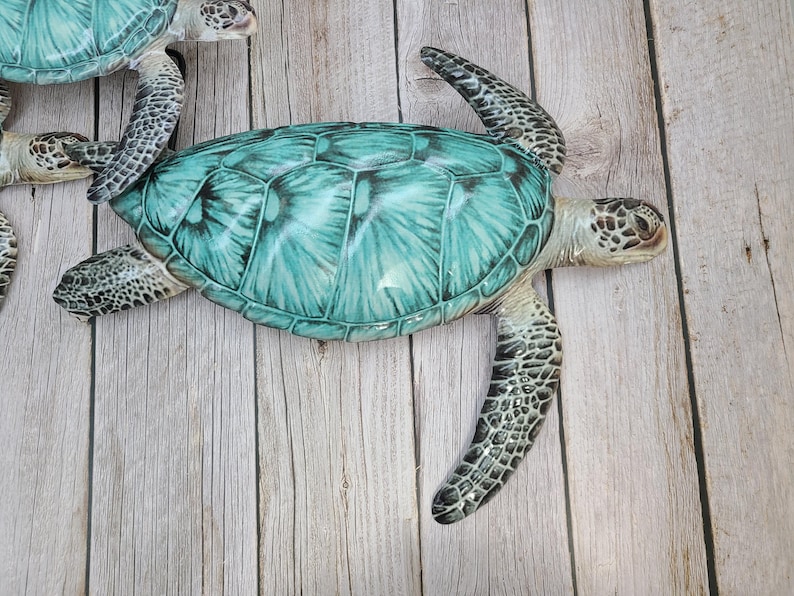 May include: Several decorative sea turtles with turquoise and green shells and speckled flippers are arranged on a light wood-grain surface. The turtles have detailed features and a realistic appearance, suggesting they are decorative items.
