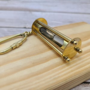 May include: A small, brass-colored hourglass keychain. The hourglass is made of glass and metal, with sand visible inside. It is attached to a brass-colored keychain ring. The item is resting on a wooden surface.