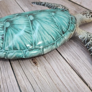 May include: A decorative sea turtle figurine with a teal and green shell and a textured, patterned head and limbs. The turtle is resting on a wooden surface.