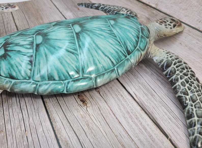 May include: A decorative sea turtle figurine with a teal and green shell and a textured, patterned head and limbs. The turtle is resting on a wooden surface.