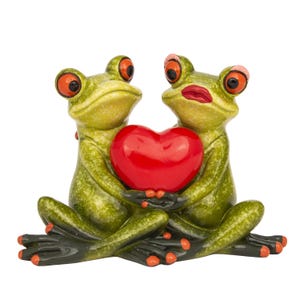 May include: A whimsical figurine featuring two green frogs holding a glossy red heart. The frogs have large orange eyes, red lips, and are posed in a loving embrace. The figurine is set against a white background.