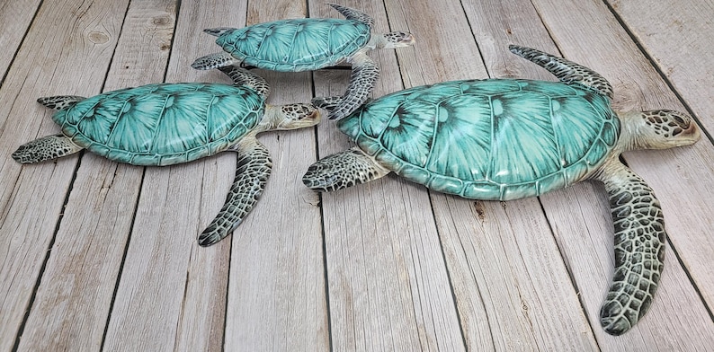 May include: Three decorative sea turtles with turquoise shells and detailed patterns are arranged on a light wood surface. The turtles vary in size, with the largest measuring approximately 20 inches in length. The turtles have mottled gray and black limbs and heads.