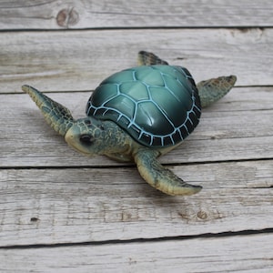 May include: A green and blue sea turtle figurine with a detailed shell and flippers. The turtle is sitting on a light brown wooden surface.