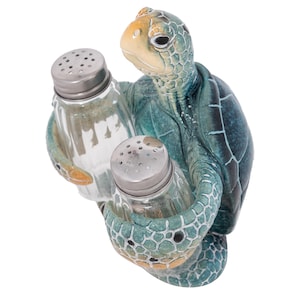 Puede incluir: Un juego de salero y pimentero de cerámica en forma de tortuga marina. La tortuga sostiene dos saleros de vidrio transparente con tapas plateadas.