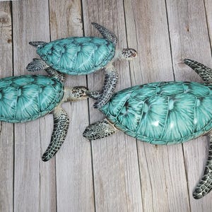 May include: Three decorative sea turtles with teal shells and speckled flippers are arranged on a light wood-grain surface. The turtles vary in size, with the largest one in the foreground. The shells have a detailed, textured appearance.