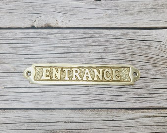 May include: A brass sign with the word "ENTRANCE" engraved on it. The sign is rectangular and has rounded corners. It is on a wooden background.
