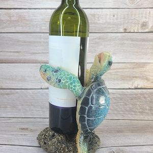May include: A decorative wine bottle holder featuring a sea turtle design. The turtle, with a blue and green shell, appears to be holding a wine bottle. The base is textured and the bottle has a blank label.