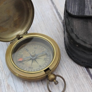 May include: A brass compass with a black leather case. The compass face has a white background with black markings and a red needle.