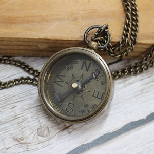 May include: A vintage-style compass pendant with a bronze-colored chain. The compass face features directional markings and a needle pointing north. The pendant is round and has a classic, antique aesthetic.