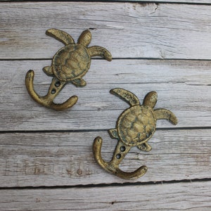 May include: Two bronze-colored metal wall hooks. Each hook features a turtle design at the top and an anchor-shaped hook below. The hooks are mounted on a light-colored, weathered wooden surface.