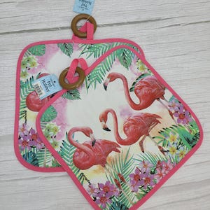 May include: Two square pot holders with a pink flamingo design. The pot holders have a white background with pink flamingos, tropical flowers, and green leaves. They have a pink border and a wooden ring for hanging.