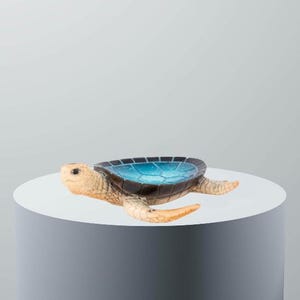 Peut inclure: Une figurine de tortue en céramique avec une carapace bleue et noire et un corps brun clair. La tortue est couchée sur le ventre, la tête tournée sur le côté. La tortue est sur une surface blanche.