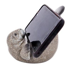 May include: A gray and brown manatee figurine holding a black smartphone in a vertical position. The figurine is designed to be a phone stand.