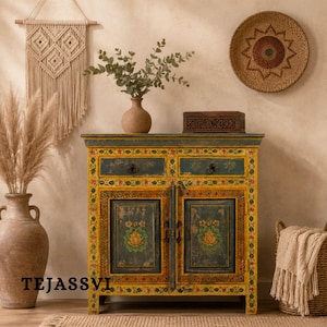 May include: A hand-painted wooden cabinet with two doors and two drawers. The cabinet is yellow with green and red floral accents. The doors feature a lotus flower design. A vase with greenery sits on top. The word "TEJASSVI" is visible.