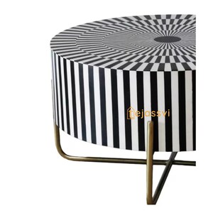 Op de afbeelding: Een ronde bijzettafel met een zwart-wit geometrisch patroon. Het tafelblad heeft een concentrisch cirkelontwerp, terwijl de zijkanten verticale strepen hebben. De tafel wordt ondersteund door goudkleurige metalen poten.