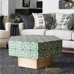 May include: A geometric patterned coffee table with a light green and white design. The table has a light wood base and is placed on a dark grey patterned rug. The table is in front of a white sofa with decorative cushions.