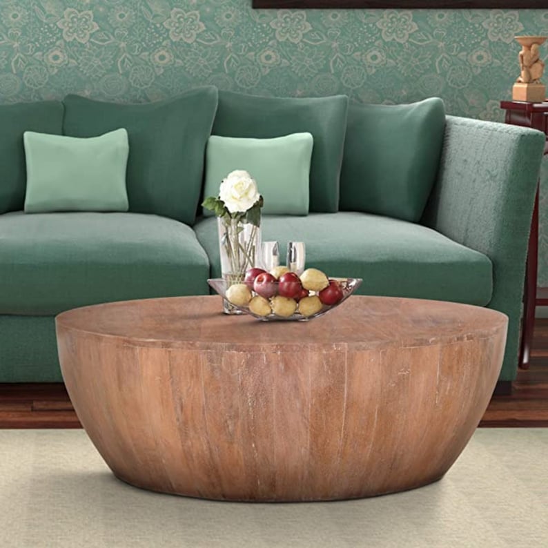 K&ouml;nnte beinhalten: Ein gro&szlig;er, runder Couchtisch aus Holz mit hellbraunem Finish. Der Tisch steht vor einem gr&uuml;nen Sofa mit hellgr&uuml;nen Kissen. Eine Glasschale mit Obst und eine Vase mit Blumen stehen auf dem Tisch.