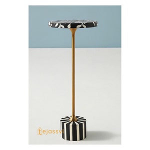 Puede incluir: Una mesa auxiliar decorativa con una tapa redonda con un estampado floral en blanco y negro. La mesa tiene un tallo dorado y una base con rayas verticales en blanco y negro. La mesa mide aproximadamente 61 cm de altura.