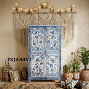 Peut inclure: Un buffet floral blanc et bleu à quatre portes. Le meuble est orné de motifs floraux bleus et de quincaillerie en laiton. Le buffet fait partie d'un décor de pièce de style bohème, avec une décoration murale en macramé, des plantes et des coussins décoratifs.