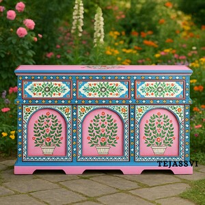 May include: A hand-painted wooden cabinet with a pink base and blue trim. The cabinet features three drawers and three arched door panels, each adorned with floral designs in white, orange, and green. The word "TEJASSVI" is visible on the right side.