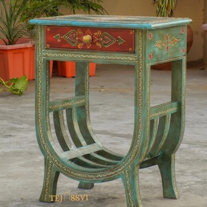 May include: A hand-painted side table with a drawer. The table is teal with gold accents and a red drawer. The drawer features a gold knob and floral design. The table has a curved magazine rack.