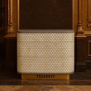 May include: A decorative chest of drawers with a gray top and a gold base. The body features a cream-colored, patterned design. The word "TEJASSVI" is printed in black on the gold base. The chest has three drawers.